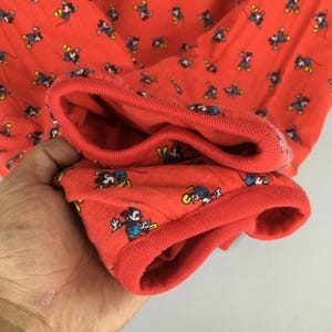 May include: Red long-sleeved shirt with a repeating pattern of Mickey Mouse in various poses. The cuffs and neckline are a solid red, and the fabric appears to be a soft knit. The shirt is likely for children or adults.