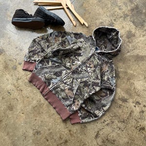 K&ouml;nnte beinhalten: Ein Camouflage-Muster-Kapuzenpullover mit Rei&szlig;verschluss und braunem Besatz, flach auf einer Betonoberfl&auml;che liegend. Schwarze Turnschuhe und Holzkleiderb&uuml;gel sind ebenfalls sichtbar. Der Hoodie hat eine Kapuze mit wei&szlig;em Besatz.