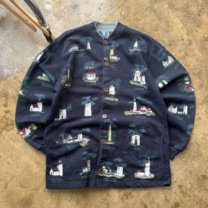 Könnte beinhalten: Marineblaue Knopfjacke mit einem sich wiederholenden Muster aus Leuchttürmen in Weiß, Rot und Gelb. Die Jacke hat einen hellblauen Kragen und braune Knöpfe. Die Leuchttürme sind vor einem dunkelgrünen und marinefarbenen Hintergrund.
