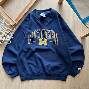 May include: A navy blue crewneck sweatshirt featuring the yellow text MICHIGAN WOLVERINES and a large yellow block M logo with the text EST. 1817. The sleeve cuff displays the yellow Lee Sport logo, and the neckline includes a triangular ribbed detail.