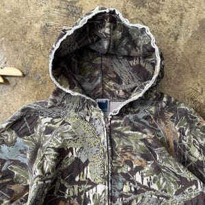 K&ouml;nnte beinhalten: Ein Kapuzenpullover mit durchgehendem Rei&szlig;verschluss in einem Tarnmuster mit Gr&uuml;n-, Braun- und Graut&ouml;nen. Die Kapuze hat eine wei&szlig;e, ausgefranste Einfassung. Das Sweatshirt hat ein Etikett mit der Aufschrift "PPFM".