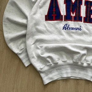May include: A white heathered sweatshirt featuring large, block-style letters with a blue and red plaid pattern that spell out part of a word. Below the large letters, the word Alumni is embroidered in blue script on the front of the garment.