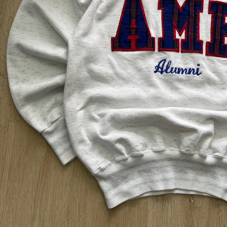 May include: A white heathered sweatshirt featuring large, block-style letters with a blue and red plaid pattern that spell out part of a word. Below the large letters, the word Alumni is embroidered in blue script on the front of the garment.