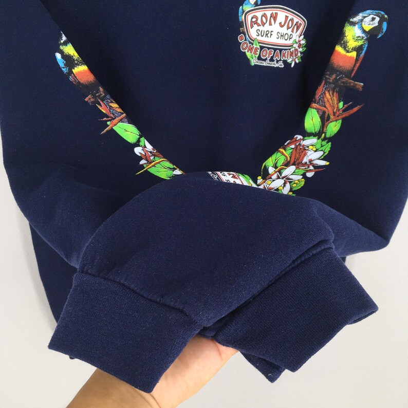 Puede incluir: Sudadera azul marino con un dise&ntilde;o colorido de loro y flores. La sudadera presenta el texto "RON JON SURF SHOP" y "ONE OF A KIND". Las mangas tienen un dise&ntilde;o de loro y flores.