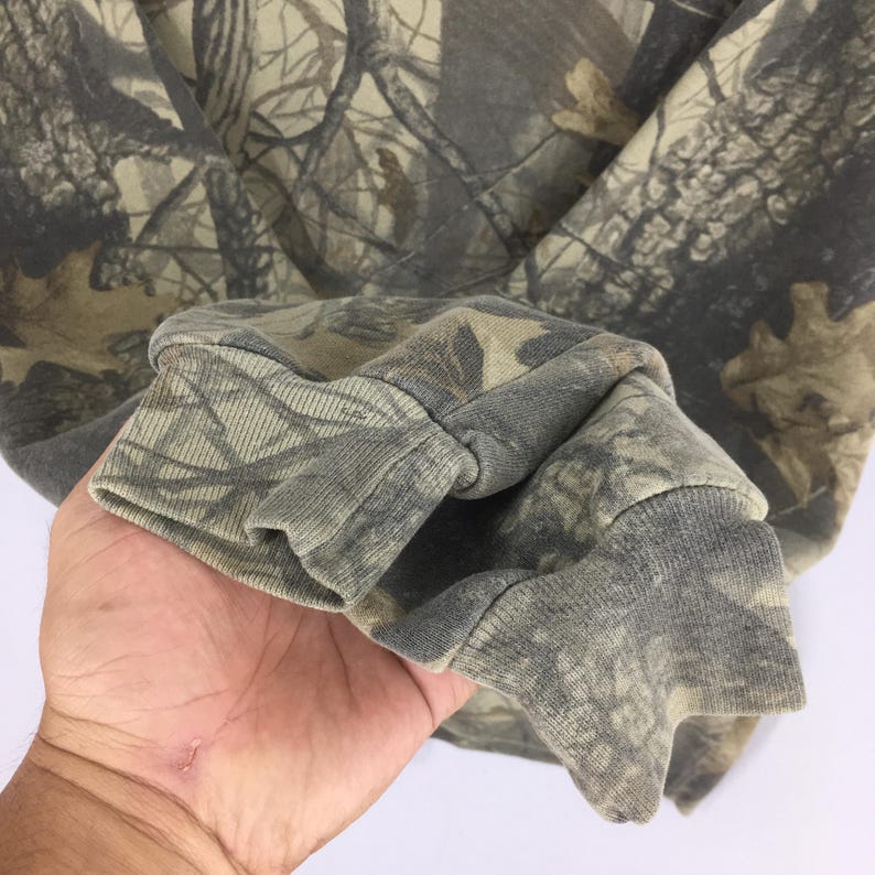 May include: Close-up of a camouflage-patterned sweatshirt sleeve. The fabric features a blend of greens, browns, and greys, mimicking a forest environment. The cuff is a solid grey-green colour.