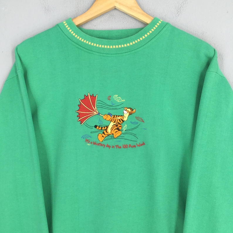 May include: Green sweatshirt featuring an embroidered image of Tigger holding a red umbrella in a windy scene. The sweatshirt has a yellow and white striped collar and the text "It's a blustery day in The 100 Acre Wood".