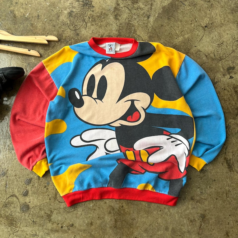 K&ouml;nnte beinhalten: Ein Vintage-Mickey-Mouse-Sweatshirt. Das Sweatshirt zeigt eine gro&szlig;e Grafik von Mickey Mouse auf blauem Hintergrund mit gelben Akzenten. Die &Auml;rmel sind rot und gelb, und der Kragen und die Manschetten sind rot.
