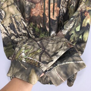 K&ouml;nnte beinhalten: Eine Tarnjacke mit durchgehendem Rei&szlig;verschluss und einem Waldmuster. Die Jacke weist eine Mischung aus Braun-, Gr&uuml;n- und Beiget&ouml;nen auf, die Bl&auml;tter und &Auml;ste nachahmen. Die Manschetten sind in einem dunkleren Farbton des Tarnmusters gehalten.