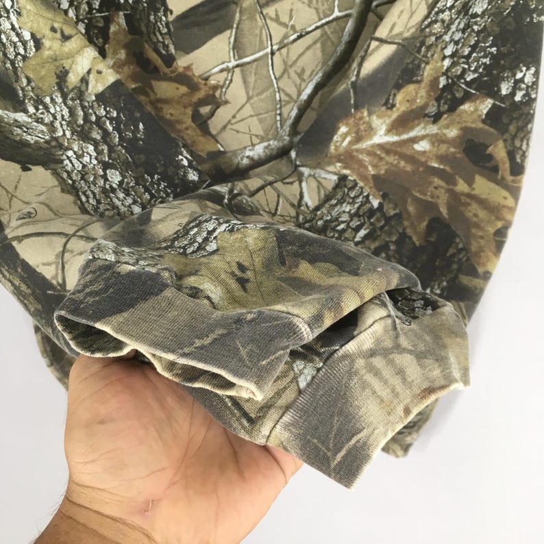 Puede incluir: Primer plano de una manga de sudadera con estampado de camuflaje. La tela presenta una mezcla de tonos marrones, beiges y verdes, imitando un entorno forestal. El pu&ntilde;o est&aacute; doblado, revelando la tela interior.