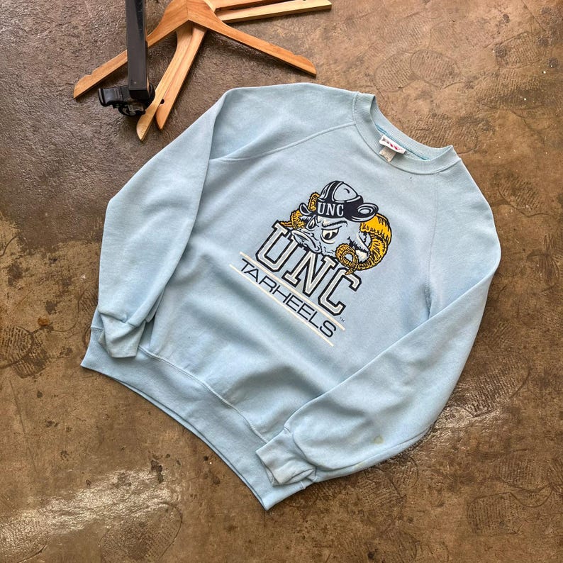 May include: A light blue crewneck sweatshirt featuring a graphic of the UNC Tarheels mascot wearing a hat. The design includes the text UNC and TARHEELS in navy blue and white lettering, with a yellow ram horn accent on the mascot illustration.