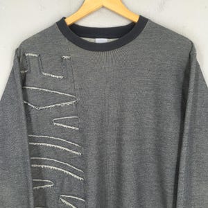 May include: A gray sweatshirt with a dark gray crew neck and a unique design on the left side. The design features white stitching that creates geometric shapes. The sweatshirt is hanging on a wooden hanger.