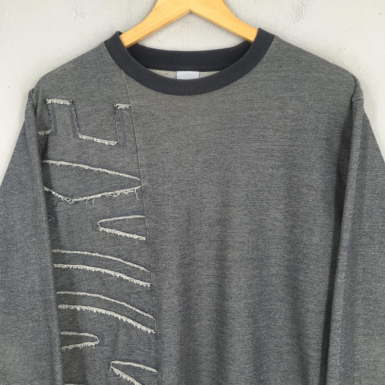 May include: A gray sweatshirt with a dark gray crew neck and a unique design on the left side. The design features white stitching that creates geometric shapes. The sweatshirt is hanging on a wooden hanger.