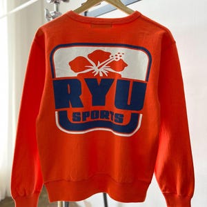May include: An orange sweatshirt with a large graphic on the back. The graphic features a white rectangle with a red and white flower, and the words "RYU SPORTS" in navy blue. The sweatshirt has long sleeves and a ribbed neckline and cuffs.