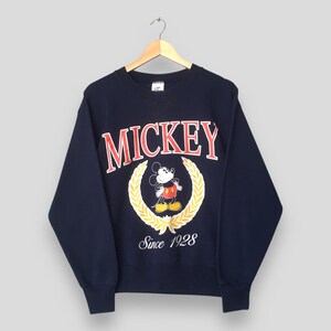 Peut inclure: Sweat-shirt bleu marine avec le mot "MICKEY" en lettres rouges au-dessus d'un dessin de Mickey Mouse et d'une couronne de laurier dorée. Le texte "Since 1928" est en dessous. Le sweat-shirt est suspendu à un cintre en bois.