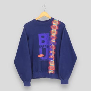 Puede incluir: Una sudadera azul marino de manga larga con un gran gráfico morado que incluye el texto BLUE FAMILY y UCB. Una franja vertical estampada en rojo, amarillo y azul claro recorre el lado derecho del pecho, mostrando formas geométricas.