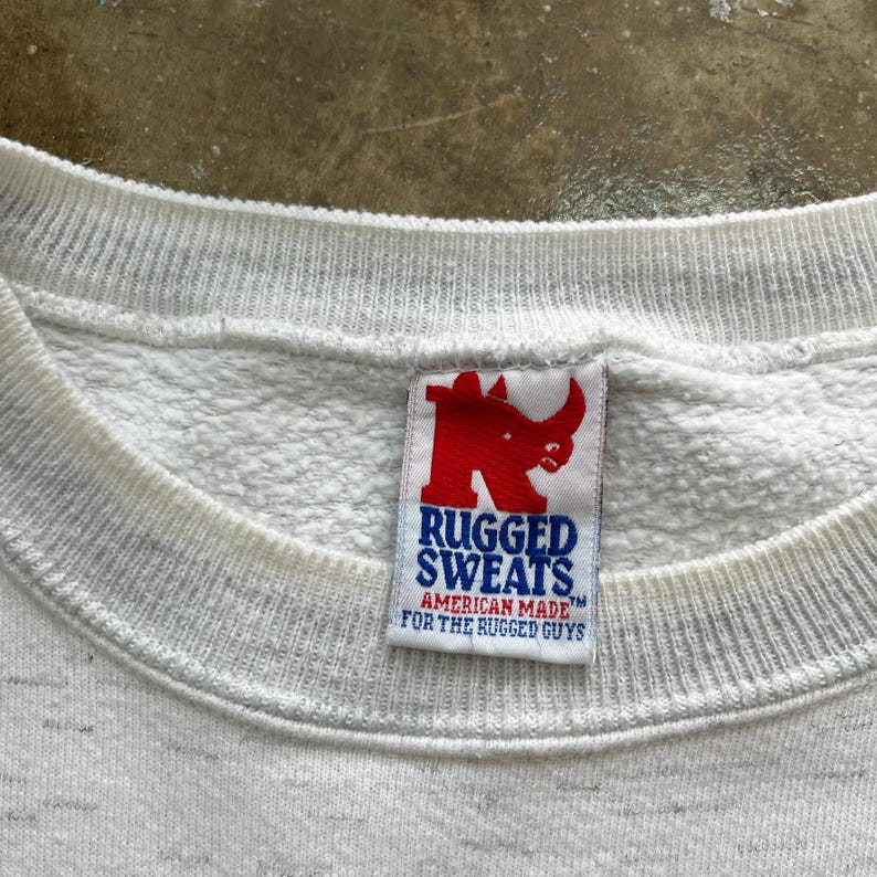 May include: A white crewneck sweatshirt with a heathered texture. The interior collar features a white fabric tag with a red rhinoceros logo and blue text that reads RUGGED SWEATS AMERICAN MADE FOR THE RUGGED GUYS.