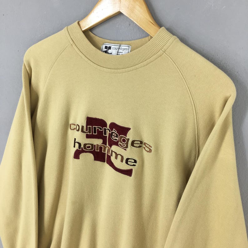 May include: A tan-colored Courr&egrave;ges homme sweatshirt with a crew neck and long sleeves. The brand name and logo are embroidered in burgundy and tan thread on the front. The sweatshirt is hanging on a wooden hanger.