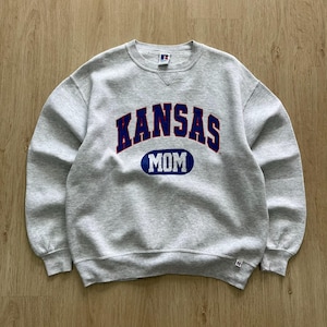 May include: A grey crew neck sweatshirt featuring the word "KANSAS" in blue and red arched lettering, and "MOM" in a blue oval. The sweatshirt has a vintage style.