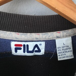May include: Close-up of a navy blue FILA sweatshirt. The white tag displays the FILA logo in red and blue. The tag also indicates the sweatshirt is size L, made of 80% cotton and 20% polyester, and made in Malaysia. The care instructions are on the back.