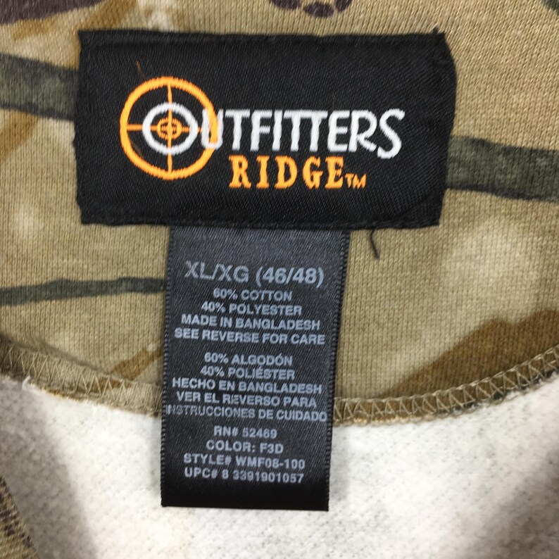May include: Close-up of a camouflage-patterned garment with a black tag. The tag features the "Outfitters Ridge" logo and size information: XL/XG (46/48). The fabric composition is 60% cotton and 40% polyester. Made in Bangladesh.