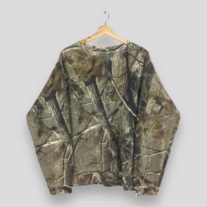 May include: A long-sleeved camouflage sweatshirt hangs from a wooden hanger. The shirt features a realistic forest pattern in shades of brown, green, and tan, with a crew neck and ribbed cuffs. The fabric appears to be a medium weight, suitable for cooler weather.