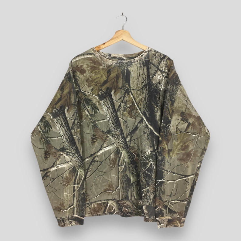 May include: A long-sleeved camouflage sweatshirt hangs from a wooden hanger. The shirt features a realistic forest pattern in shades of brown, green, and tan, with a crew neck and ribbed cuffs. The fabric appears to be a medium weight, suitable for cooler weather.