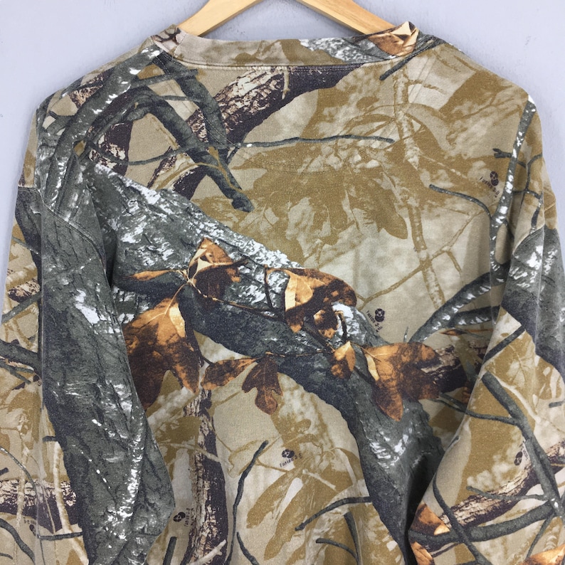 May include: A long-sleeved camouflage shirt in shades of brown, green, and tan. The pattern mimics a forest environment, with tree branches and leaves. The shirt is hanging on a wooden hanger.
