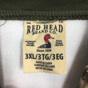 K&ouml;nnte beinhalten: Nahaufnahme eines Etiketts der Marke Red Head Brand Co. Das beige Etikett zeigt ein rotes und schwarzes Entenlogo und den Text "Since 1856". Es enth&auml;lt auch die Gr&ouml;&szlig;e "3XL/3TG/3EG" und "Made in China".