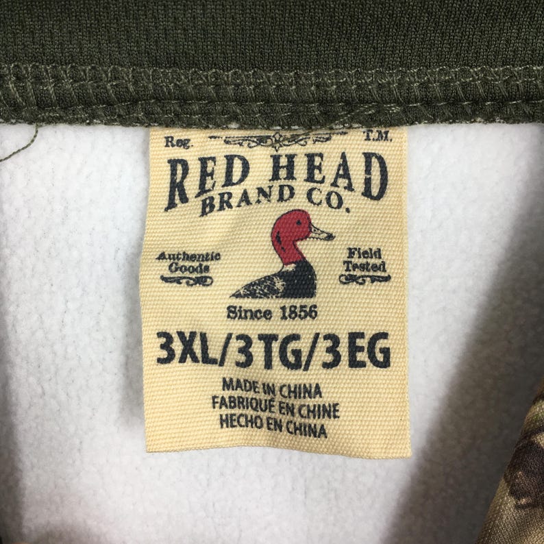 K&ouml;nnte beinhalten: Nahaufnahme eines Etiketts der Marke Red Head Brand Co. Das beige Etikett zeigt ein rotes und schwarzes Entenlogo und den Text "Since 1856". Es enth&auml;lt auch die Gr&ouml;&szlig;e "3XL/3TG/3EG" und "Made in China".