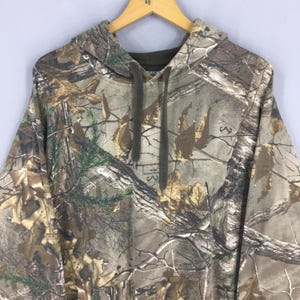 Op de afbeelding: Een hoodie met camouflagepatroon en een capuchon met trekkoord. De stof heeft een mix van bruine, groene en bruine tinten, die een bosrijke omgeving nabootsen. De hoodie hangt aan een houten kleerhanger tegen een lichtgrijze achtergrond.