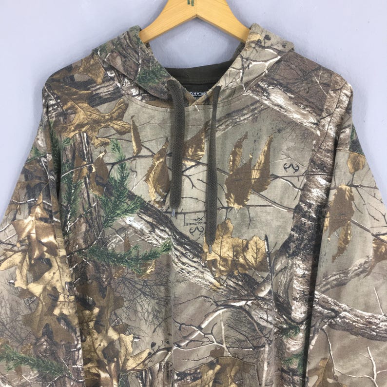 Op de afbeelding: Een hoodie met camouflagepatroon en een capuchon met trekkoord. De stof heeft een mix van bruine, groene en bruine tinten, die een bosrijke omgeving nabootsen. De hoodie hangt aan een houten kleerhanger tegen een lichtgrijze achtergrond.