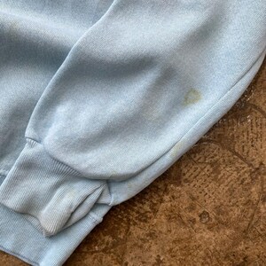 May include: A light blue long-sleeved sweatshirt featuring a ribbed cuff at the wrist. The fabric displays a soft, textured knit surface with visible faint yellowish discoloration spots located on the sleeve near the cuff area.