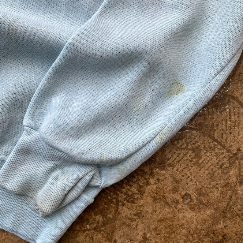 May include: A light blue long-sleeved sweatshirt featuring a ribbed cuff at the wrist. The fabric displays a soft, textured knit surface with visible faint yellowish discoloration spots located on the sleeve near the cuff area.