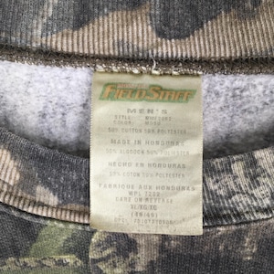 Puede incluir: Primer plano de una sudadera con estampado de camuflaje con una etiqueta. La etiqueta dice "Field Staff" e incluye detalles sobre la composici&oacute;n de la tela, que es 50% algod&oacute;n y 50% poli&eacute;ster. La etiqueta tambi&eacute;n indica que la prenda fue fabricada en Honduras.
