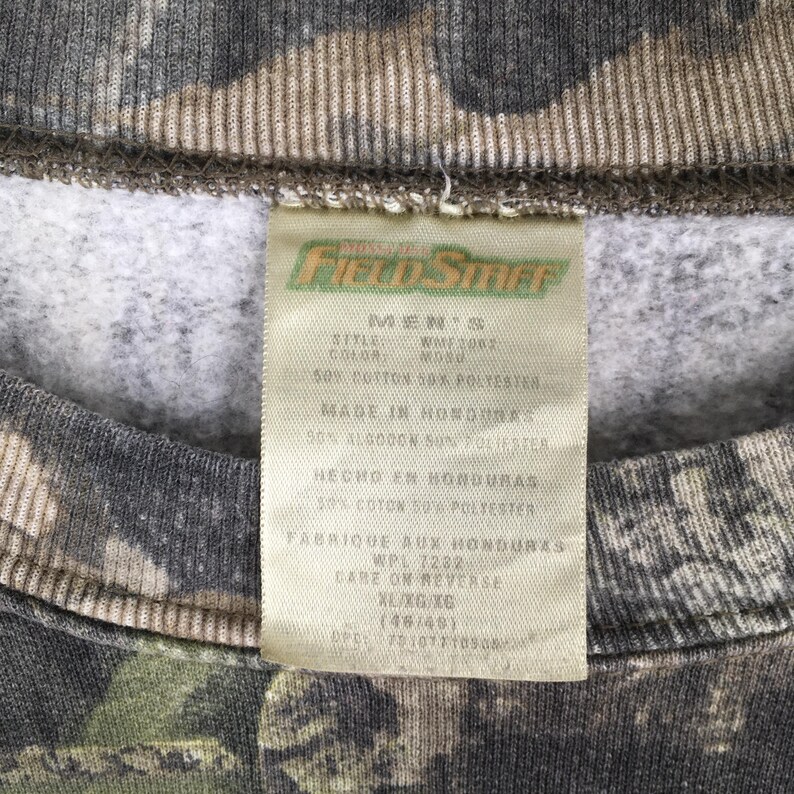Puede incluir: Primer plano de una sudadera con estampado de camuflaje con una etiqueta. La etiqueta dice "Field Staff" e incluye detalles sobre la composici&oacute;n de la tela, que es 50% algod&oacute;n y 50% poli&eacute;ster. La etiqueta tambi&eacute;n indica que la prenda fue fabricada en Honduras.