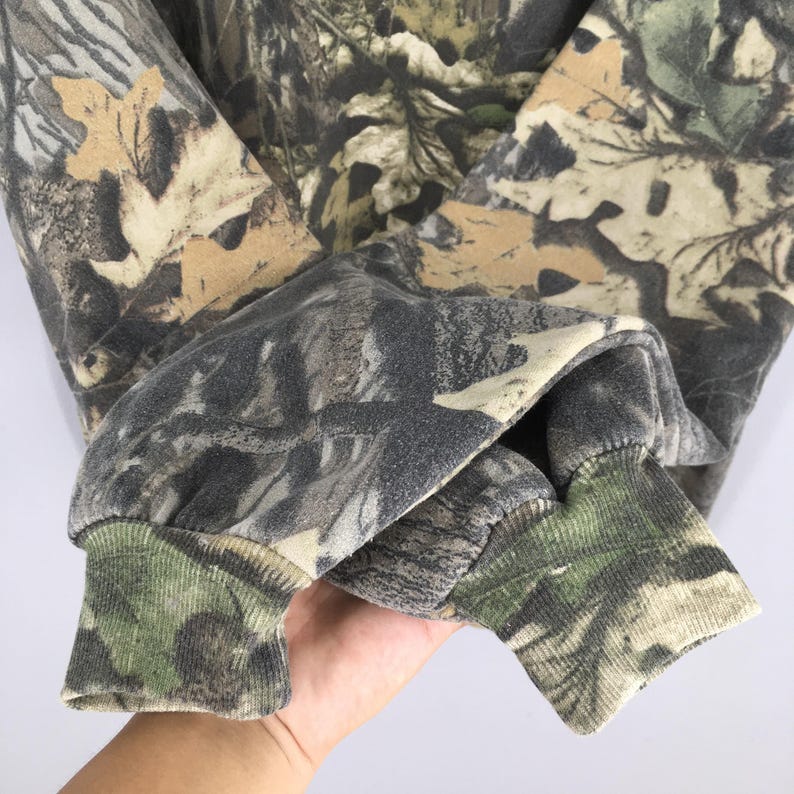 May include: A long-sleeved camouflage sweatshirt with a forest pattern. The cuffs and sleeves are visible, showcasing the blend of greens, browns, and tans. The fabric appears to be a soft, comfortable material, suitable for outdoor activities.