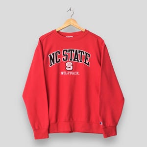 May include: A red NC State Wolfpack sweatshirt with the words "NC STATE" in white block letters with a black outline. The letter "S" is in the center, and the word "WOLFPACK" is below. The sweatshirt is hanging on a wooden hanger.