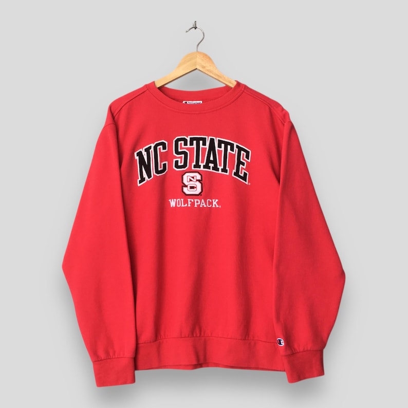 May include: A red NC State Wolfpack sweatshirt with the words "NC STATE" in white block letters with a black outline. The letter "S" is in the center, and the word "WOLFPACK" is below. The sweatshirt is hanging on a wooden hanger.