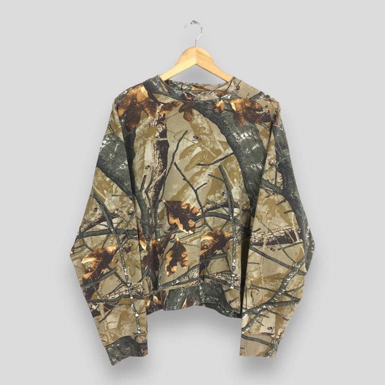 May include: Long-sleeved camouflage shirt with a crew neck, hanging on a wooden hanger. The shirt features a pattern of brown, green, and tan branches and leaves, creating a woodland camouflage design. The fabric appears to be a medium-weight material.