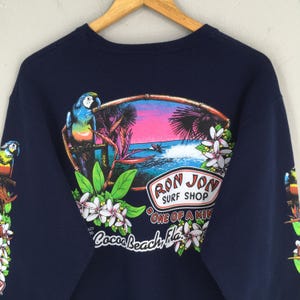 Puede incluir: Sudadera azul marino con un dise&ntilde;o tropical. Presenta un loro, palmeras y una escena oce&aacute;nica. El gr&aacute;fico incluye el texto "Ron Jon Surf Shop" y "Cocoa Beach, Fla.". Flores blancas y hojas verdes enmarcan el dise&ntilde;o central.