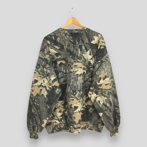 May include: A long-sleeved sweatshirt with a camouflage pattern featuring shades of brown, green, and tan. The design incorporates leaf and tree bark elements, creating a woodland aesthetic. The sweatshirt is hanging on a wooden hanger.