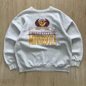 May include: A gray crewneck sweatshirt featuring the words "UNIVERSITY OF MINNESOTA" in yellow and maroon lettering. Above the text is a maroon and yellow graphic of a gopher mascot and the words "GOLDEN GOPHERS".