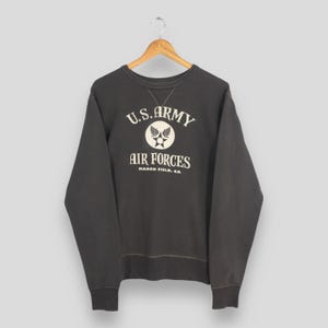May include: A charcoal gray long-sleeve sweatshirt featuring a cream-colored graphic print that reads U.S. ARMY AIR FORCES MARCH FIELD, CA with a central star and wing emblem. The garment has a crew neckline and ribbed cuffs and hem.