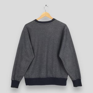 May include: A gray sweatshirt with a navy blue collar, cuffs, and hem. The sweatshirt is hanging on a wooden hanger against a plain background. The back of the sweatshirt is visible.