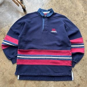 May include: A navy blue rugby shirt with a denim collar and red and white striped accents. The shirt features the Canterbury of New Zealand logo and a quarter-zip closure. The sleeves have red and white stripes.