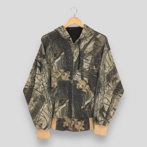 May include: A camouflage hooded zip-up jacket. The jacket features a woodland camouflage pattern in shades of brown, green, and tan. It has a black interior lining and tan-colored cuffs and waistband. The jacket is hanging on a wooden hanger.
