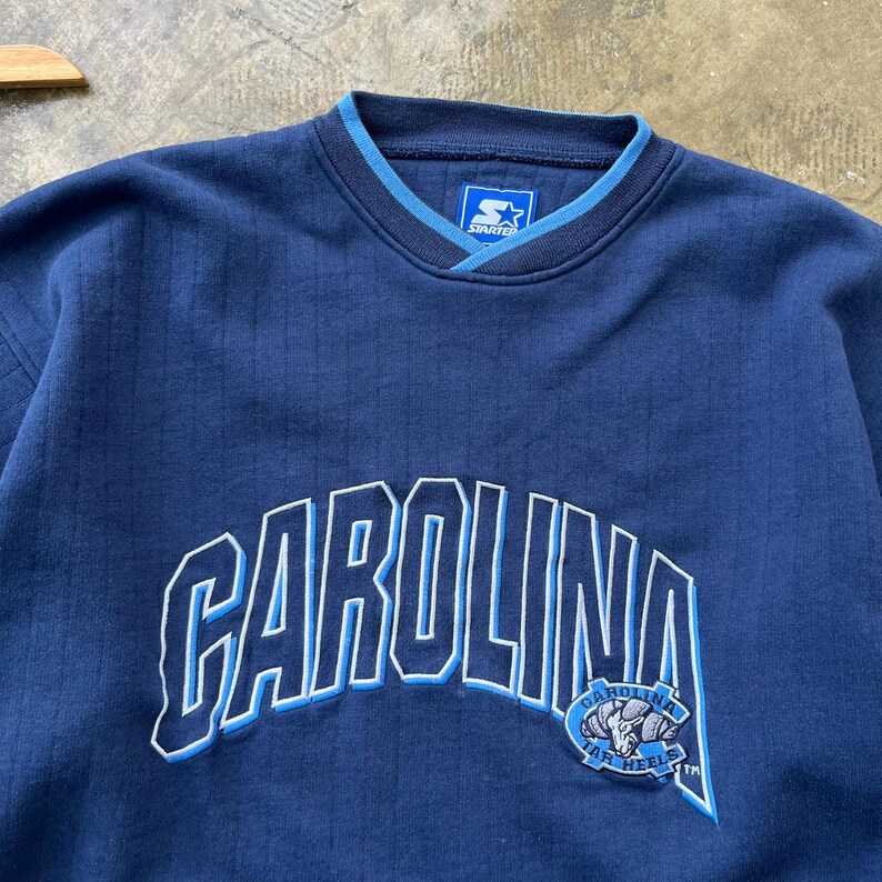 May include: A navy blue long-sleeve sweatshirt with a ribbed texture. The front features the word CAROLINA in large white and light blue embroidered letters, with a Tar Heels logo patch and a Starter brand label at the inner neckline.