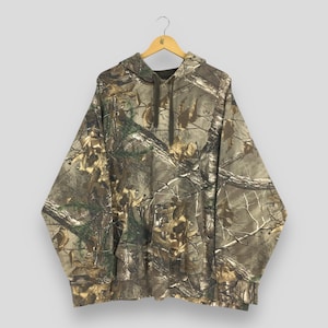 Op de afbeelding: Een camouflage hoodie met een capuchon met trekkoord en een kangoeroezak. Het patroon heeft tinten bruin, groen en beige, die lijken op een bosrijke omgeving. De hoodie hangt aan een houten hanger.