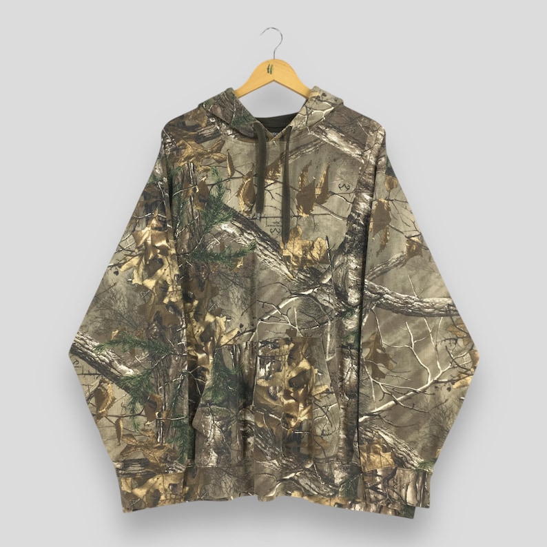 Op de afbeelding: Een camouflage hoodie met een capuchon met trekkoord en een kangoeroezak. Het patroon heeft tinten bruin, groen en beige, die lijken op een bosrijke omgeving. De hoodie hangt aan een houten hanger.