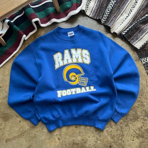 May include: A royal blue crewneck sweatshirt featuring the word RAMS in white block letters above a yellow and white football helmet graphic, with the word FOOTBALL printed in yellow block letters below the helmet on the front of the garment.